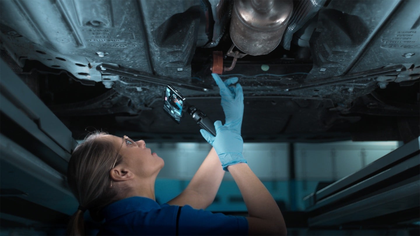 A Ford technician inspects the underside of a vehicle using a specialized tool.